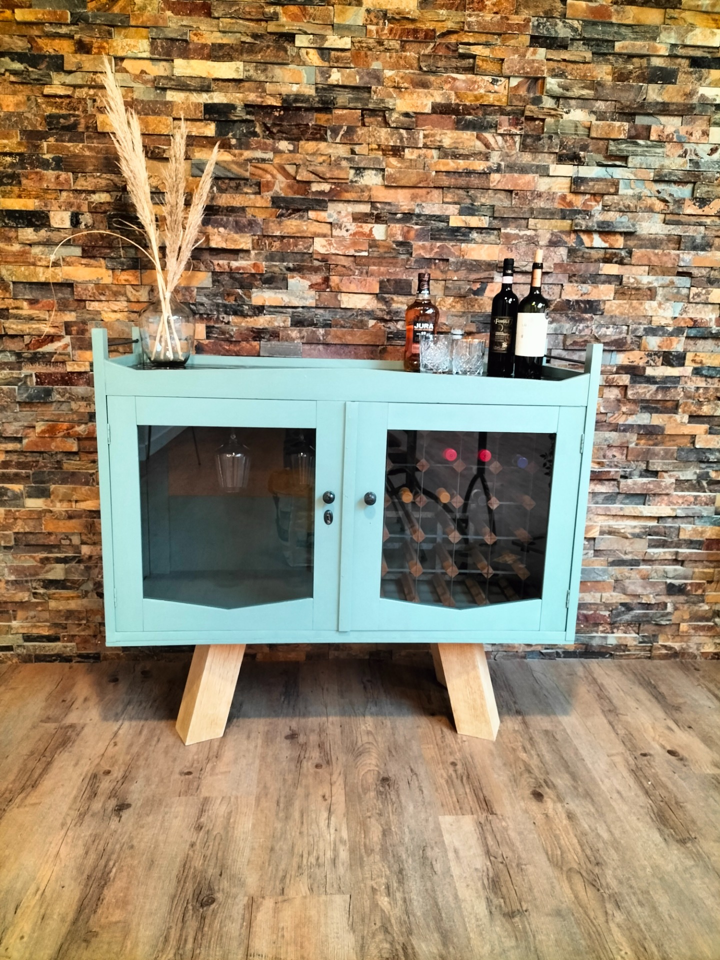 Witte vitrinekast met glazen deuren, flessen en glazen op plank, tegen kleurrijke stenen muur. Interieur. Studio aankleding. Originele kast.