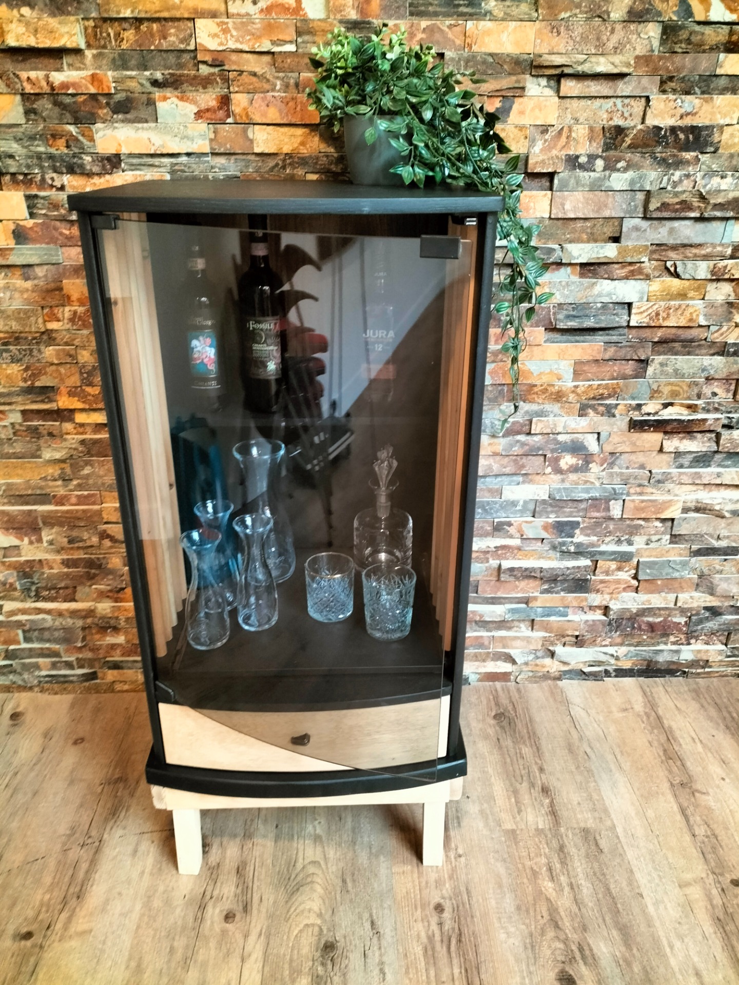 Glazen vitrine met flessen en glazen, tegen stenen muur met groene plant bovenin. Vintage. Drankkast. Studio inrichting. Interieur. Podcast studio.